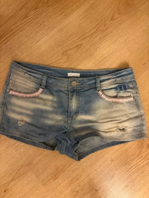 Blå jeansshorts med rosa detaljer - Säljer ett par ljusblå, lågmidjade jeansshorts med slitningar och coola rosa detaljer vid fickorna. Shortsen har klassisk femficksdesign, bälteshällor och knappstängning. På de två sista bilderna syns rostfläckar som tyvärr inte går bort, men de syns knappt om man inte lyfter på de rosa detaljerna. Storlek 42, men passar som S eller XS.