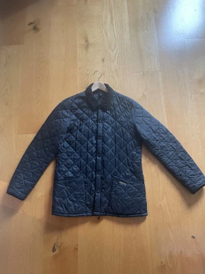 Mörkblå quiltad vindjacka med krage - Stilren mörkblå vindjacka från Barbour med quiltat mönster och klassisk krage. Jackan har tryckknappar framtill, två fickor och raka ärmar. Materialet är lätt och smidigt, perfekt för blåsiga dagar. Passar dig som gillar enkel och tidlös stil.