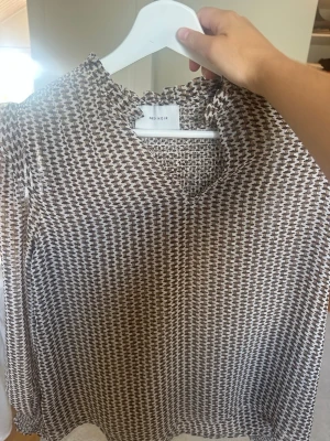 Mönstrad blus från Neo Noir - Säljer en snygg blus från Neo Noir med brunt och vitt grafiskt mönster. Blusen har långärmad design, lätt rynkad krage och v-ringning. Tillverkad i ett tunt, luftigt material som känns skönt mot huden. Perfekt att styla med jeans eller kjol.
