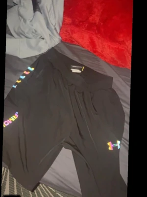 Svarta shorts från Under Armour - Säljer ett par svarta shorts från Under Armour med färgglada loggor på benet och sidan. De har en elastisk midja och är perfekta för träning eller chill. Materialet känns lätt och andas bra.