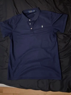 Marinblå pikétröja från Polo Ralph Lauren - Klassisk marinblå pikétröja från Polo Ralph Lauren med vit broderad logga på bröstet. Tröjan har korta ärmar, krage och knappar framtill. Perfekt till både jeans och shorts för en stilren look.