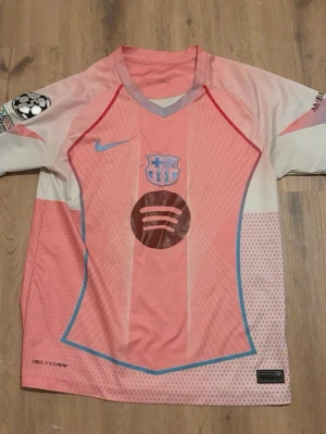 Rosa FC Barcelona matchtröja Nike - Säljer en rosa och vit FC Barcelona matchtröja med blå detaljer och tryck. Tröjan har klubbmärke, Nike-logga och sponsortryck på framsidan samt Lamine Yamal och nummer 19 på ryggen. Champions League-märke på ärmen och UNHCR-logga bak. Perfekt för fotbollsfans!