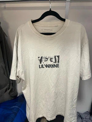 Vit Lil Wayne t-shirt - Säljer en vit lil Wayne merch t-shirt med ett svart tryck av Lil Wayne på bröstet. T-shirten har rund hals och korta ärmar. Perfekt för dig som gillar streetwear och vill ha en unik tee med artistmotiv.