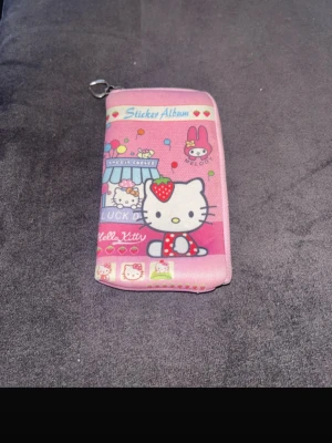 Rosa Hello Kitty plånbok - Supergullig rosa plånbok med Hello Kitty-motiv och söta detaljer som nallar och ballonger. Plånboken har dragkedja runtom och färgglada tryck med texten 'Sticker Album'. Perfekt för dig som älskar kawaii och vill ha något unikt i väskan.