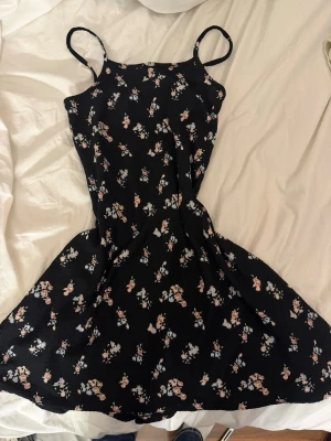 Svart blommig sommarklänning - Söt svart klänning med smala axelband och blommigt mönster i rosa och vitt. Klänningen har en utställd kjol och är perfekt för varma dagar. Tillverkad i ett lätt och mjukt material som känns skönt mot huden.