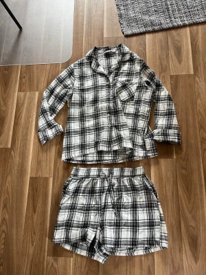Rutiga pyjamas i svartvitt - Säljer en mysig pyjamas med svartvitt rutigt mönster. Setet består av en långärmad skjorta med knappar och bröstficka samt matchande shorts. Perfekt för chillkvällar hemma. Materialet är mjukt och luftigt.
