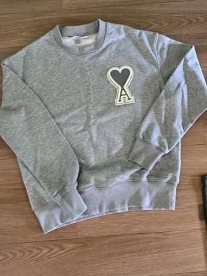 Grå ami sweatshirt  - Säljer en stilren grå sweatshirt med rund hals och ribbade muddar. På bröstet finns en stor applikation med ett hjärta och bokstaven A i vitt och grått. Perfekt för dig som gillar enkel men snygg design.