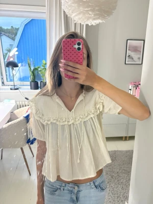 krämvit blus med spets och volang - Söt krämvit blus med korta puffärmar, volang och spetsdetaljer över bröstet. Blusen har stickat material upp till och runt där nere, knytband vid halsen.
