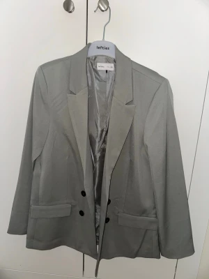 Ljusgrå kavaj från Lefties - Stilren ljusgrå kavaj från Lefties med klassisk passform, två framfickor och dubbla knappar. Kavajen har slät finish och helfodrad insida, perfekt för en snygg och clean look.