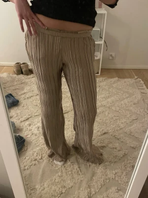 Beige plisserade  byxor - Säljer ett par beige byxor med plisserad struktur och raka ben. Byxorna har resår i midjan och är tillverkade i ett mjukt, flowy material som ger en avslappnad vibe. Perfekta för chill dagar eller när du vill vara bekväm men ändå se snygg ut. Alrig använda lappen finns kvar