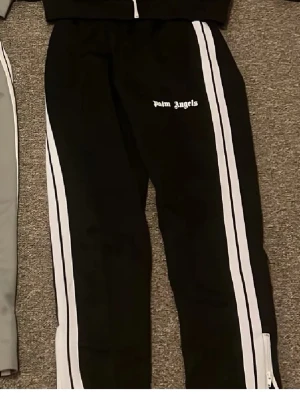 Palm Angels mjukisbyxor med ränder - Palm Angels mjukisbyxor i svart och grått med vita sidoränder och logga på vänster lår. Byxorna har resår i midjan och dragkedja vid benslut. Perfekta för en avslappnad streetwear-look.