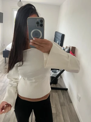 Vit ribbad topp med vida ärmar - Trendig vit topp med ribbad midja och vida, långa ärmar. Toppen har en något croppad passform och en stilren, enkel design som passar perfekt till jeans eller kjol. Materialet känns mjukt och stretchigt.
