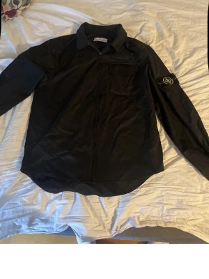 Svart overshirt från Stone Island - Säljer en svart overshirt från Stone Island med dragkedja framtill och klassisk patch på vänster ärm. Skjortan har en bröstficka och är tillverkad i ett lätt, glansigt material som ger en clean look. Perfekt för lager-på-lager och streetwear vibes.
