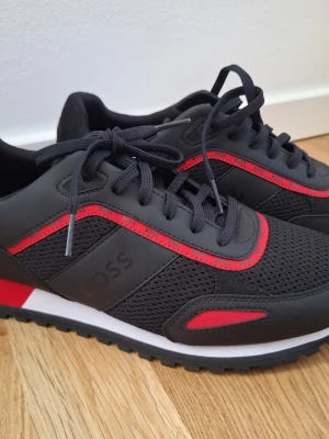 Svarta och röda sneakers från BOSS - Nya, oanvända! Snygga sneakers från BOSS i svart mesh och syntet med röda detaljer och logga på sidan. Skorna har svarta snören, vit och röd sula samt sportig design. Perfekta för dig som vill ha en modern och stilren look.
