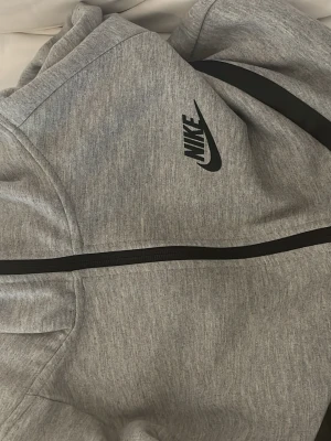 Grå zip hoodie från Nike - Snygg grå hoodie från Nike med svart dragkedja och logga på bröstet. Hoodien har huva och är tillverkad i mjukt sweatshirtmaterial. Perfekt för en sportig och avslappnad stil.