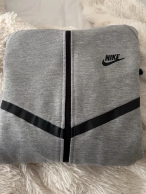Grå Nike hoodie med dragkedja - Snygg grå hoodie från Nike med hel dragkedja framtill och svart Nike-logga på bröstet. Hoodien har svarta detaljer i form av en diagonal linje över bröstet och en huva med dragsko. Perfekt för en sportig och avslappnad stil. Kontakta för intresse/frågor!