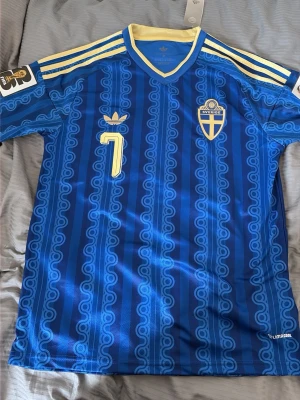 Blå Sverige fotbollströja Adidas - Säljer en blå fotbollströja från Adidas med Sveriges landslagsemblem på bröstet och nummer 7 i gult. Tröjan har gula detaljer vid krage och axlar samt ett mönster i olika blå nyanser. Tillverkad i lätt och ventilerande material, perfekt för träning eller match.