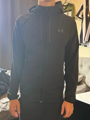 Sällsynt Svart vindjacka från Under Armour - Snygg svart vindjacka från Under Armour med dragkedja framtill och justerbar huva. Jackan har diskret logga på bröstet och är tillverkad i ett lätt, vindtätt material. Perfekt för dig som vill ha en stilren och funktionell jacka.
