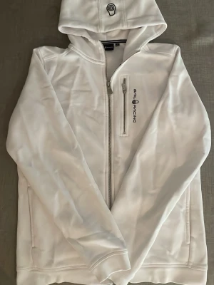Vit hoodie från Sail Racing - Säljer en fräsch vit hoodie från Sail Racing med dragkedja framtill och en extra ficka med zip på bröstet - Nytt pris ca 1000kr. Hoodien har en broderad logga på huvan och på bröstet. Perfekt för dig som gillar stilren och sportig look.