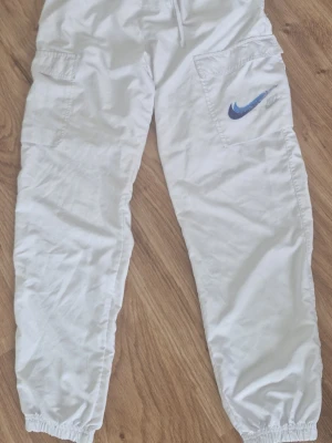 Nike byxor - Säljer ett par nya vita cargobyxor från Nike med stora fickor på sidorna och elastiska benslut. Byxorna har en blå Nike-logga på ena benet och är perfekta för en avslappnad och sportig stil.