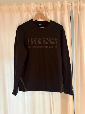 Svart långärmad tröja från BOSS - Snygg svart långärmad tröja från BOSS med diskret logga i mönstrad design på bröstet. Tröjan har rund halsringning och dekorativa detaljer vid sidorna. Perfekt för en stilren och avslappnad look.