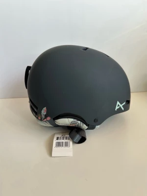 HELT NY svart snowboardhjälm - Anon Greta - storlek M (57-58cm) - Helt ny svart / grå Anon Greta snowboardhjälm i storlek M (57-59 cm). Tidlös stilren modell i mattsvart / grått med mönstrat foder i turkos och korall. Justerbara remmar och praktiska geometriska vädrings-perforeringar. Kvalitativ hjälm för snowboard, skidåkning eller andra utomhusaktiviteter. Levereras med instruktionsmanual och HELT NY i originalförpackning. Nypris 999:-.