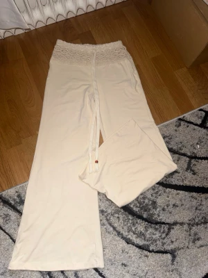 Beiga vida byxor med spetsdetalj - Säljer ett par beiga, vida byxor med hög midja och bred spetskant upptill. Byxorna har dragsko i midjan och är gjorda i ett mjukt, stretchigt material som känns riktigt skönt mot huden. Perfekta för chill dagar hemma eller när du vill ha något bekvämt. En av kulorna på snöret saknas annars i perfekt skick, passar kortare 