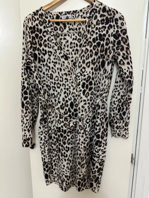 Leopardmönstrad omlottklänning KappAhl - Snygg leopardmönstrad omlottklänning från KappAhl i storlek 38. Klänningen har långa ärmar, knyt i midjan och v-ringad hals. Perfekt för dig som vill sticka ut med ett djurmönster och en bekväm passform.