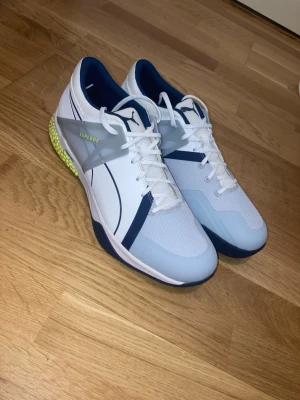 Puma Explode Skor - Säljer ett par nya vita Puma Explode inomhusskor med marinblå och grå detaljer. Skorna har vita snören och blå/gul sula. Perfekta för sport och träning, men funkar också som vardagliga skor.