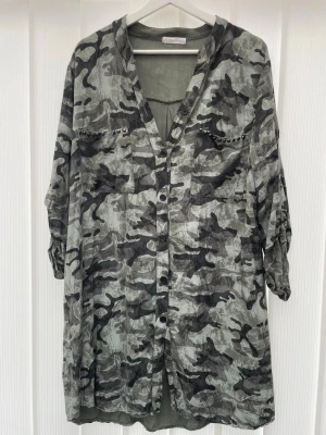 Grå camouflageklänning med knappar - Säljer en loose fit klänning i grått camouflage-mönster med knappar framtill och v-ringning. Klänningen har trekvartsärm och bröstfickor med små nitar som detalj. Perfekt för dig som gillar en avslappnad och cool stil.