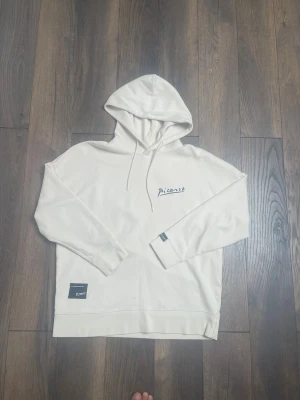 Beige hoodie med Picasso-tryck - Snygg beige hoodie med huva och dragsnören. Framsidan har en liten broderad text 'Picasso' och på ryggen finns ett stort konstinspirerat tryck i svartvitt. Mjuk och skön bomullskänsla, perfekt för en avslappnad stil.