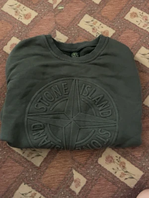 Mörkgrön Stone Island sweatshirt - Snygg mörkgrön sweatshirt från Stone Island med stort broderat logotypmärke på bröstet. Tröjan har rund halsringning och ribbade muddar. Perfekt för dig som gillar streetwear och vill ha en stilren look.