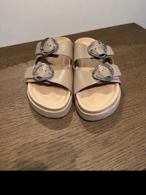 Sweeks sandaler - Snygga beige sandaler med två breda remmar och stora, runda silverfärgade spännen. Sandalerna har en chunky sula och öppen tå. Tillverkade i ett slätt material som ger en clean look. Perfekta för dig som gillar minimalistisk stil.