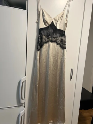 Champagnefärgad klänning med spets - Långklänning i champagnefärgad satin med tunna axelband och svart spetsdetalj över bysten.Den har en glansig finish och en elegant, feminin siluett som ger en lyxig känsla. Perfekt för dig som vill ha något extra fint.