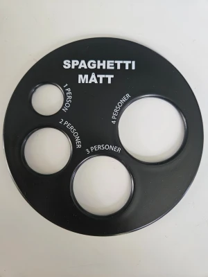 Svart spaghettimått i plast - Praktiskt svart spaghettimått i plast med fyra olika hål för att mäta portioner till 1, 2, 3 eller 4 personer. Perfekt för att få rätt mängd pasta varje gång. Rund form och enkel att använda i köket.