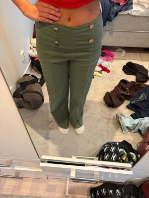 Gröna byxor med guldknappar - Snygga gröna byxor med hög midja och raka ben. Fyra dekorativa guldknappar framtill ger en unik touch. Byxorna har en clean look utan synliga fickor och är perfekta för dig som vill sticka ut lite extra.