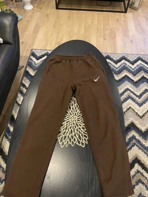 Nike - Säljer ett par bruna mjukisbyxor från Nike med klassisk vit logga på vänster ben. Byxorna har resår i midjan och raka ben, perfekta för chill dagar. Tillverkade i mjukt material som känns skönt mot huden. Enkla att matcha med hoodie eller t-shirt.