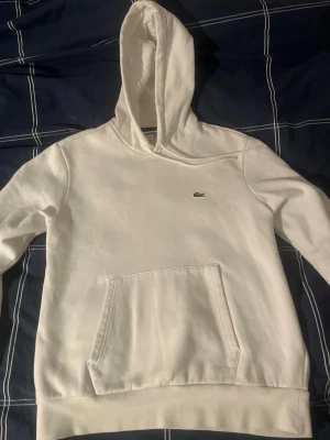 Lacoste Hoodie - Säljer en clean vit hoodie från Lacoste med klassisk krokodil-logga på bröstet. Hoodien har stor ficka framtill och en skön huva med dragsko. Perfekt för dig som gillar stilrena plagg med street-vibe.