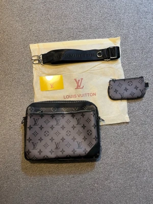 Trio bag - Snygg axelväska från Louis Vuitton i grått och svart med klassiskt monogrammönster. Väskan har justerbar axelrem, flera fack med dragkedja och matchande liten pouch. Tillverkad i canvas med detaljer i läder.