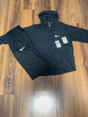 Nike Miller - Snyggt svart träningsset från Nike med matchande jacka och byxor. Jackan har huva och dragkedja framtill samt Nike-logga på bröstet. Byxorna har resår i midjan och vit Nike-logga på benet. Perfekt för träning eller chill. Materialet är lätt och andas.