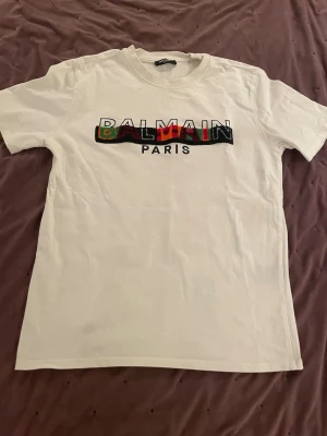 Vit Balmain Paris t-shirt - Vit t-shirt från Balmain Paris med färgglatt broderad logga på bröstet. Klassisk passform med rund hals och korta ärmar. Perfekt för dig som vill sticka ut med en ikonisk streetstyle-look.