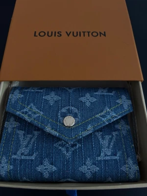 Blå denimplånbok  - Snygg blå plånbok i denim med ikoniskt monogrammönster och gul kontrastsöm. Plånboken har en triangulär klaff med silverfärgad knapp och flera kortfack på insidan. Perfekt accessoar för dig som gillar exklusiv och trendig design.