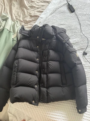 Moncler jacka - Snygg svart dunjacka från Moncler med puffig design och huva. Jackan har tryckknappar, dragkedja och en ficka med Moncler-logga på ärmen. Perfekt för kalla vinterdagar och riktigt trendig look.