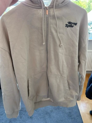 Beige zip hoodie med tryck - Säljer en beige hoodie med dragkedja och huva. På bröstet finns ett svart tryck med texten 'Strong Together'. Hoodien har två fickor framtill och ribbade muddar vid ärmar och nederkant. Perfekt för en chill och avslappnad stil.