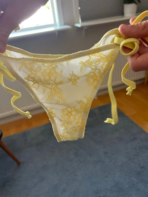 Gula spets-trosor med knytband SHEIN - Säljer ett par gula trosor från SHEIN i storlek S. Trosorna har tunna knytband i sidorna och är gjorda i mesh med blommig spetsdetalj. Materialet är lätt och genomskinligt, perfekt för dig som gillar en feminin och trendig stil.
