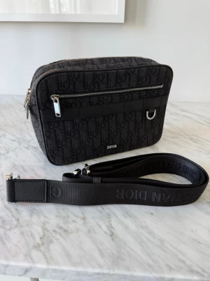 Svart axelväska CD - Snygg svart axelväska med monogrammönster och justerbar axelrem. Väskan är i väldigt bra skick förutom att den e lite nopprig! lite solbränd vid framsidan syns inte så mycket!