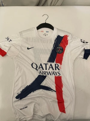 Vit PSG matchtröja med tryck - Säljer en vit PSG matchtröja från Nike med Qatar Airways-tryck, klubbmärke och färgstarka penseldrag i blått och rött. Nummer 7 och namnet Kvartskhelia på ryggen. Tröjan har korta ärmar och Champions League-märke på ena ärmen. Inte spelar edition.