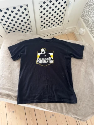 Svart t-shirt Evig Kapten AIK - Svart t-shirt med tryck av Nils-Eric Johansson och texten 'Evig Kapten' i vitt och gult. Motiv med AIK-tema på bröstet. Klassisk passform och rund hals. Perfekt för dig som vill hylla en AIK-ikon. Väldigt unik och sällsynt T-shirt som ej går att få tag på längre.