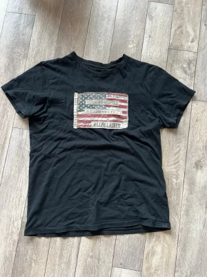 Denim & supply x Ralph Lauren T-shirt  - Snygg svart t-shirt från Ralph Lauren med amerikansk flagga och logga tryckt på bröstet. Klassisk passform och rund halsringning, perfekt till jeans eller shorts. T-shirten är i mjuk bomull och har korta ärmar.