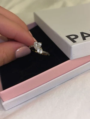 Guldfärgad ring med stenar från Pandora - Vacker guldfärgad promise ring från Pandora med två stora, gnistrande stenar i hjärt former framtill och små klara stenar längs halva ringen. Perfekt för dig som vill ha något som sticker ut och känns lyxigt..